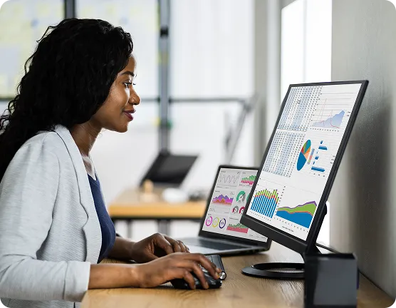 A confident businesswoman smiles as she analyses OrderEazi's comprehensive dashboard on dual monitors.