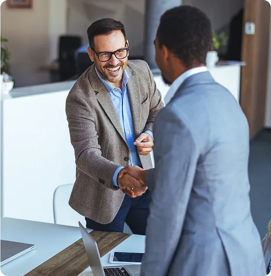 Two businessmen shake hands enthusiastically in an office setting.
