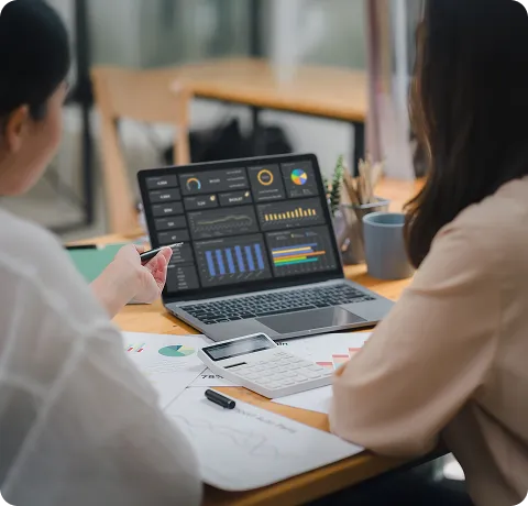 Two people sitting at a table reviewing financial documents and data charts, with a laptop displaying various colourful business analytics dashboards, a calculator, and a tablet on the table.