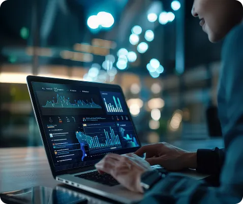 A person working on a laptop displaying detailed data analytics dashboards with charts and graphs, set in a modern workspace with soft lighting.